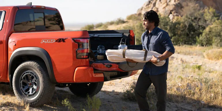 2025 Nissan Frontier PRO-4X with an open tailgate, featuring a man unloading camping gear, emphasizing its utility and versatility.
