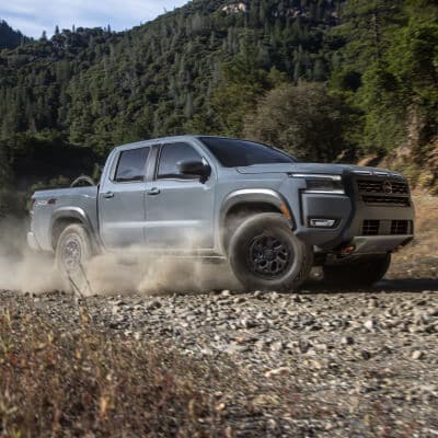 2025 Nissan Frontier driving on a rocky trail surrounded by greenery, showcasing its off-road capability and rugged design.