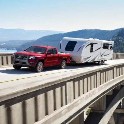 2025 Nissan Frontier towing a large travel trailer across a scenic bridge with mountains and a lake in the background, demonstrating its towing power.